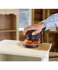 Badass 18V Cordless Orbital Sander With Killer Dust Control And Super Smooth Softgrip Handle For Hassle Free Wood Projects 12,000 Opms Compact Orange Design used on a wooden surface.
