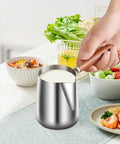 Person holding Stainless Steel Milk Warmer And Frother Pot With Wooden Handle Barista Grade Multipurpose Milk Frother Cup For Home Or Cafe 900ml Capacity Great For Latte Art Coffee Tea And Melting Chocolate