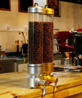 Sick Wall Mount Coffee Bean Dispenser Heavy Hitter Acrylic Canister with silver and gold finish holding coffee beans