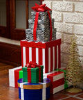 Stack of colorful wrapped gift boxes on floor beside white brick wall, with small decorative tree nearby