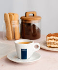 Espresso in a Lavazza cup on a table with cake, breadsticks, and a jar of coffee in the background