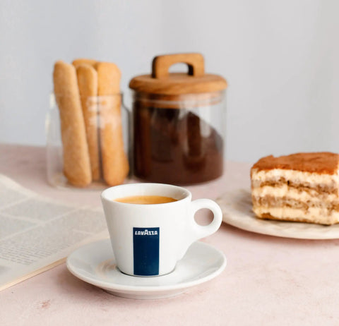Espresso in a Lavazza cup on a table with cake, breadsticks, and a jar of coffee in the background