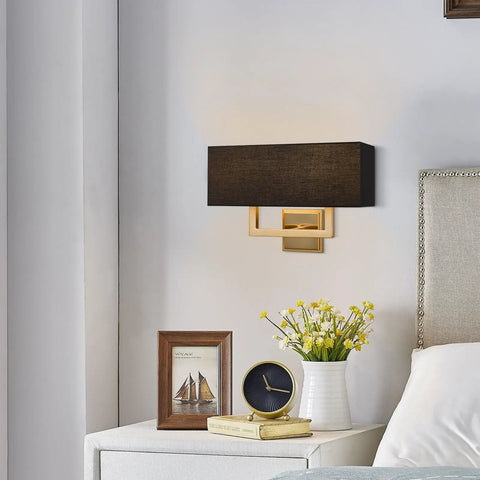 Sleek Mid Century Modern 16 Inch Two Light Wall Sconce In Cool Brass Gold Finish With Black Fabric Shade Above a Bedside Table