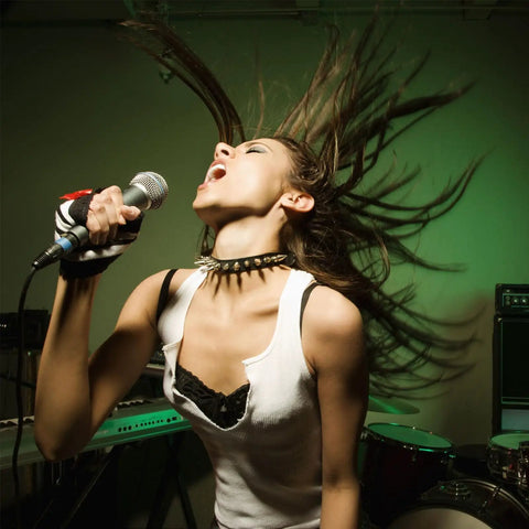 Woman singing energetically into a microphone with hair flowing, wearing a spiked choker and a white top in a music studio.