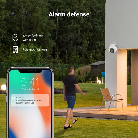Dual Lens 1080P Outdoor Security Camera With Color Night Vision, Active Light And Siren Alarm, PIR Motion Detection, mounted on a house wall with a young man receiving alerts on his phone.