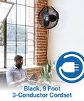 18 Inch Industrial Wall Mount Fan With Pivoting Head For Directional Airflow One Sixth Horsepower Powder Coated Steel Black Great For Garage Or Workshop mounted on a brick wall above a man using a laptop.