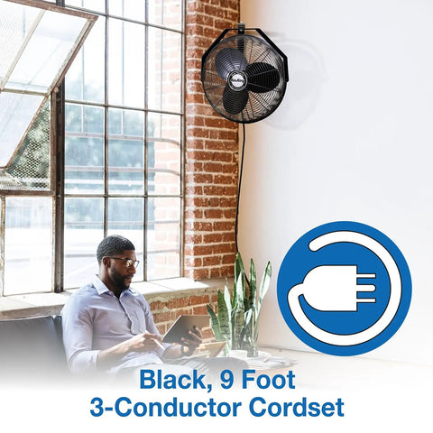 18 Inch Industrial Wall Mount Fan With Pivoting Head For Directional Airflow One Sixth Horsepower Powder Coated Steel Black Great For Garage Or Workshop mounted on a brick wall above a man using a laptop.