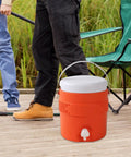 Portable Sports Drink Dispenser With Heavy Insulation Keeps Backyard Faves Stay Frosty All Weekend And Sip Smooth At Tailgates While Hanging Out In Vibrant Orange placed on wooden deck by fishing chairs.