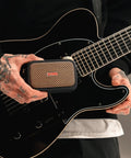 Person holding Ultra Portable 5W Smart Guitar Amp With Bluetooth Speaker Headphone Amp And Mobile App For Electric Acoustic Or Bass Guitars 33 Amps 43 Effects Custom Presets 50,000 ToneCloud Tones next to a guitar