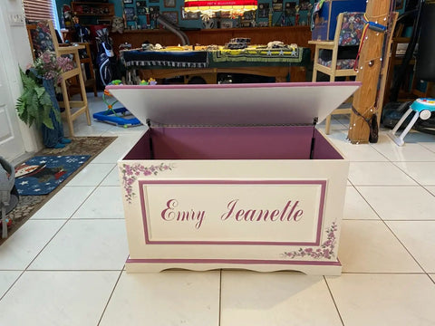 Open wooden toy chest with decorative floral design and personalized name, showing interior supported by soft close hinges for safety.