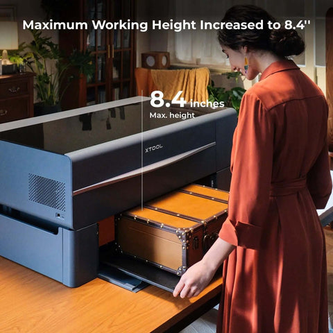 xTool P2 CO2 laser cutter with riser base; black unit on desk, brown suitcase on lower tray, 8.4-inch max height.