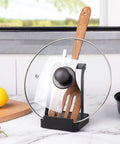 Hella Convenient Multi Function Kitchen Utensil And Pot Lid Organizer Pad With Spoon Rest Ladle Holder Catch Mat Drip Tray Washable Heavy Duty Stick Mount Black holding pot lid and spatula on a kitchen counter.