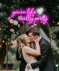 Couple kissing under neon sign reading "you're like really pretty" with greenery and flowers, creating a romantic atmosphere