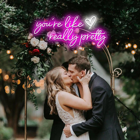 Couple kissing under neon sign reading "you're like really pretty" with greenery and flowers, creating a romantic atmosphere