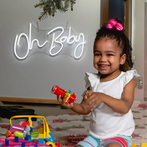Neon Sign For Wall Decor With Dimmable Brightness And Reusable Light Up Message For Baby Shower, Birthday Party, And Wedding Vibe, Cool White Glow, Large 23.5 By 11.6 Inches Neon Accent For Any Festive Space glowing 'Oh Baby' above smiling child indoors.