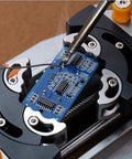 Desktop Mini Vise Dope For Soldering And DIY Projects With Adjustable Grip And Built In Lighting Magnifying Glass Attachment Smoke Exhaust Fan Ideal For Electronics Fine Crafting Repairs And Delicate Assemblies securing a blue PCB