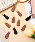 Zipper Pull Tabs Replacement Kit For Luggage Backpacks Jackets Purses And Handbags Grab And Go Easy Clip On PU Leather in black, khaki, and red-brown on a table surrounded by bags.