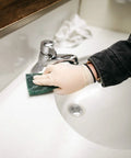 Ivory latex disposable glove used to scrub a white bathroom sink with a green scrub pad.
