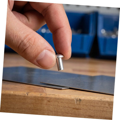 Hand holding a silver aluminum brazier head rivet M5x10 millimeters above metal sheet for leather crafting.