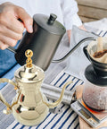 Golden stainless steel Arabic coffee pot and pour over teapot set with infuser next to coffee brewing scene on kitchen table.