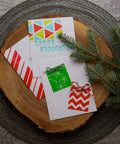 Christmas cards with geometric and striped designs on a wooden disk, accented by pine branches
