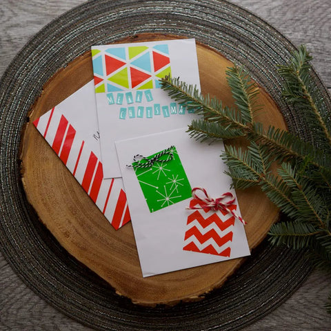Christmas cards with geometric and striped designs on a wooden disk, accented by pine branches