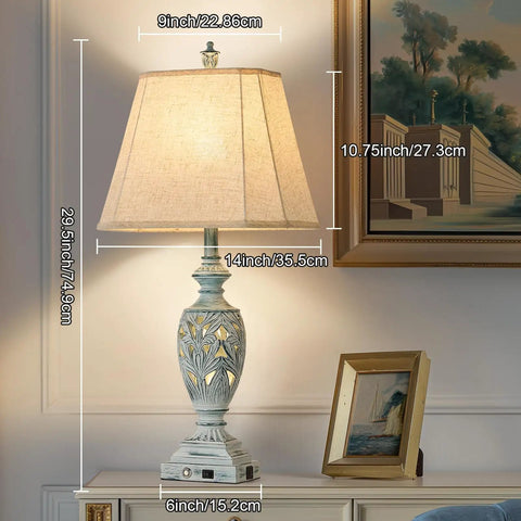 Tall decorative table lamp with a square linen shade and intricate base, placed on a nightstand.