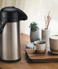 85 Oz Insulated Airpot Coffee Carafe with Pump, Stainless Steel Beverage Dispenser on wooden table with coffee cups
