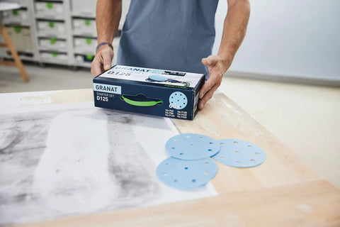 Person holding a box of Granat D125 GR Set Sanding Discs for VOC Varnishes and Hard Substrates, three discs on table