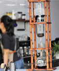 Big Batch Cold Brew Coffee Maker With Adjustable Stainless Steel Dripper And Roomy Borosilicate Glass Carafe displayed on a wooden stand.