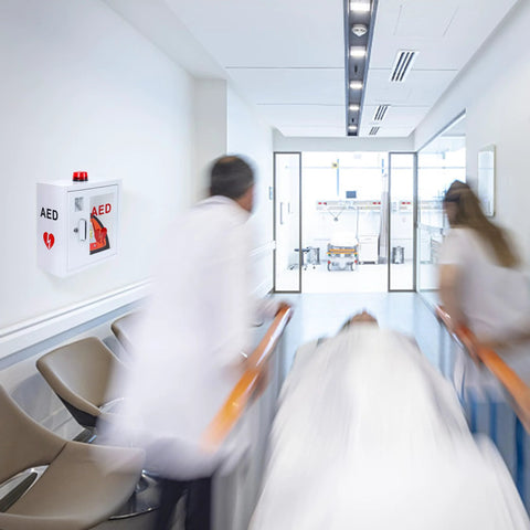 AED wall-mounted cabinet in a hospital hallway; white metal box with red alarm beacon and AED label
