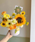 Hand holding bouquet of sunflowers, roses, and daisies in a white vase with greeting card attached.