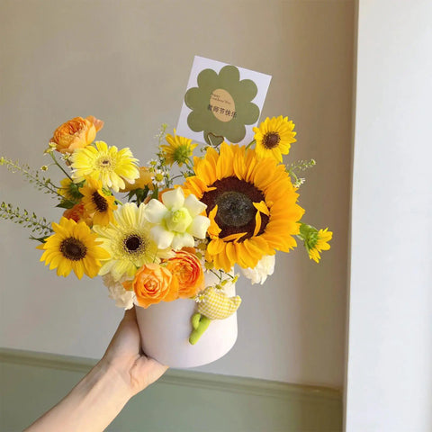 Hand holding bouquet of sunflowers, roses, and daisies in a white vase with greeting card attached.