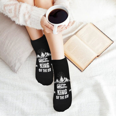 Person wearing black socks with 'King of the RV' print, holding a coffee cup; 5 Pairs Ankle Socks For Running, Gym And RV Camping - Moisture-Wicking, Quick-Dry Athletic Socks With Breathable Mesh, Snug Fit, Odor Control, No-Sweat Comfort nearby book on a cozy blanket.