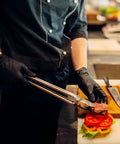 Front view: Schneider black vinyl exam gloves worn by a chef, handling food with tongs over a burger.