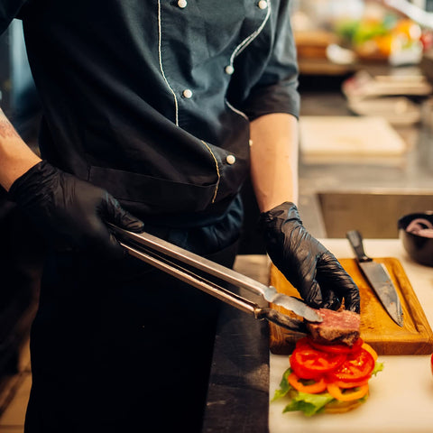 Black disposable gloves worn by a chef using tongs to handle meat on a cutting board with tomato slices nearby