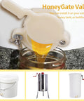 Close-up of a beige plastic honey gate valve for 5-gallon bucket, dispensing honey into a glass jar.