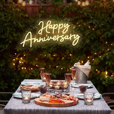 Outdoor anniversary dinner setup with Happy Anniversary LED neon sign and candle-lit table, evening garden ambiance.