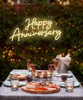 Happy Anniversary neon sign glowing over a romantic outdoor table setting with candles and drinks.