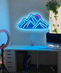 Blue neon mountain wall decor above a desk with computer, ring light, and potted plant in a modern workspace.