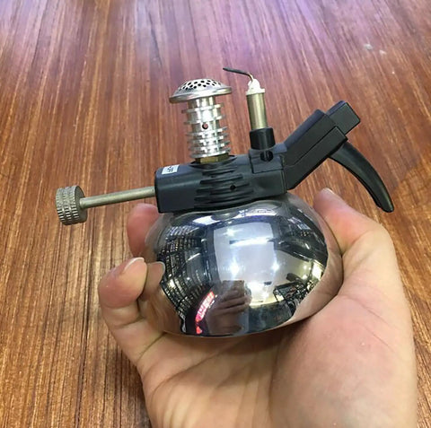 Hand holding Mini Butane Burner Portable Gas Stove With Aluminum Rack For Outdoor Camping And Home Science Experiments, 1300 C Heating, No Hoses Needed, Durable And Travel Ready, Ideal For Coffee Heating And Quick Lab Tasks.