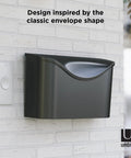 Postino Wall Mount Mailbox In Brushed Stainless Steel With Hinged Lid, Weatherproof And Dope Envelope-Inspired Front Door Mailbox for modern homes on a white brick wall.