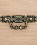 Close-up of a vintage antique brass bail pull on a wooden surface; Six Pack Vintage Antique Brass Bail Pulls For Drawers Cabinets Dressers Cupboards Set Featuring 4.1 Inch Length And 1.5 Inch Height Inter Hole Spacing 3.5 Inches Made From Zinc Alloy With Distressed Finish Retro Rustic Hardware