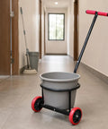 Heavy Duty Manual Transport Cart With Circular Upper Support And Open Frame Base For Easy Bucket Trolley Moving Of Loaded Containers, Two Wheel Hand Truck Carrier, Black Carbon Steel, Ideal For Cleaning Routes And Events. Bucket cart in hallway with gray bucket and red wheels