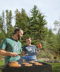 People cooking on a Park Style Charcoal Grill For Camping And Backyard Outdoor Cooking Portable Patio Barbecue For Parks And Camp Grilling 20.5 By 20.5 By 8.25 Inches Durable No Fuss Setup outdoors in a forested area.