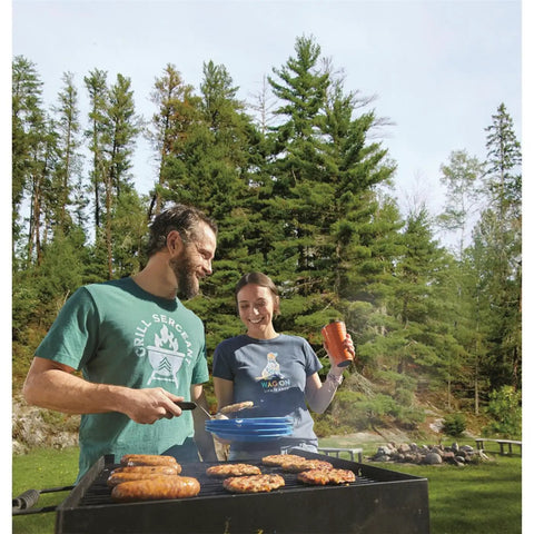 People cooking on a Park Style Charcoal Grill For Camping And Backyard Outdoor Cooking Portable Patio Barbecue For Parks And Camp Grilling 20.5 By 20.5 By 8.25 Inches Durable No Fuss Setup outdoors in a forested area.