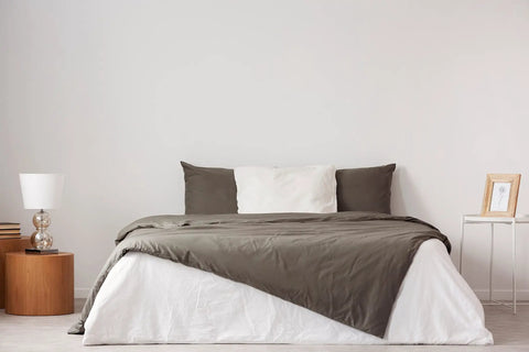 Modern bedroom with grey and white bedding, wooden side table, white lamp, and minimal decor in a cozy setting.