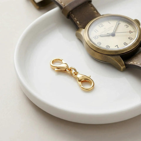 Gold double end lobster clasp, part of 20 Pcs Double End Lobster Clasp Set For DIY Jewelry Making, Necklace Extender And Bracelet Extender Clips In Gold And Silver, Zinc Alloy Construction, Replacement Clasps For Women, Great For DIY Bling And Built To Last, displayed on a white dish next to a wristwatch.