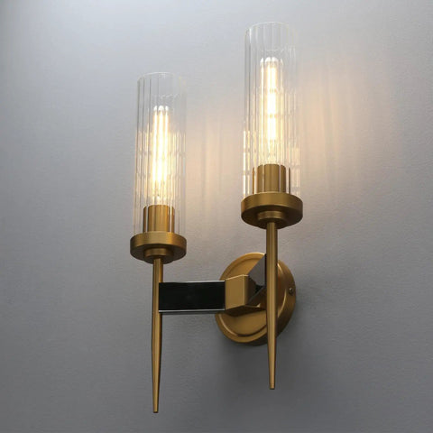 Two-Light Gold Brass Wall Sconces With 2.8 Inch Cylindrical Clear Glass Shades illuminated on a gray wall, dimmable, E26 base compatible.