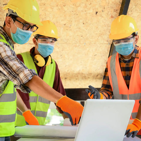 Three construction workers in orange-black heat resistant gloves reviewing plans on a laptop.